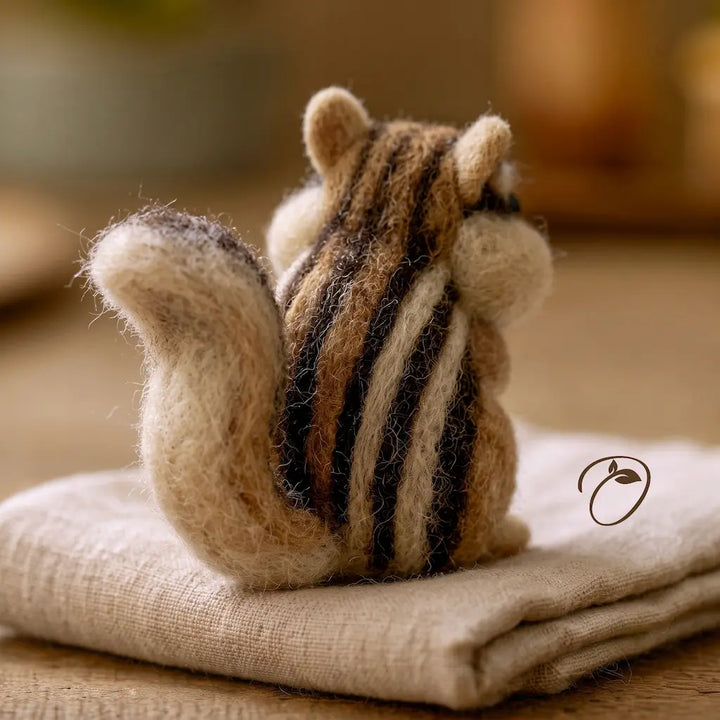 A handcrafted felted wool chipmunk figurine, shown from behind, sitting on a folded beige linen cloth on a wooden surface.