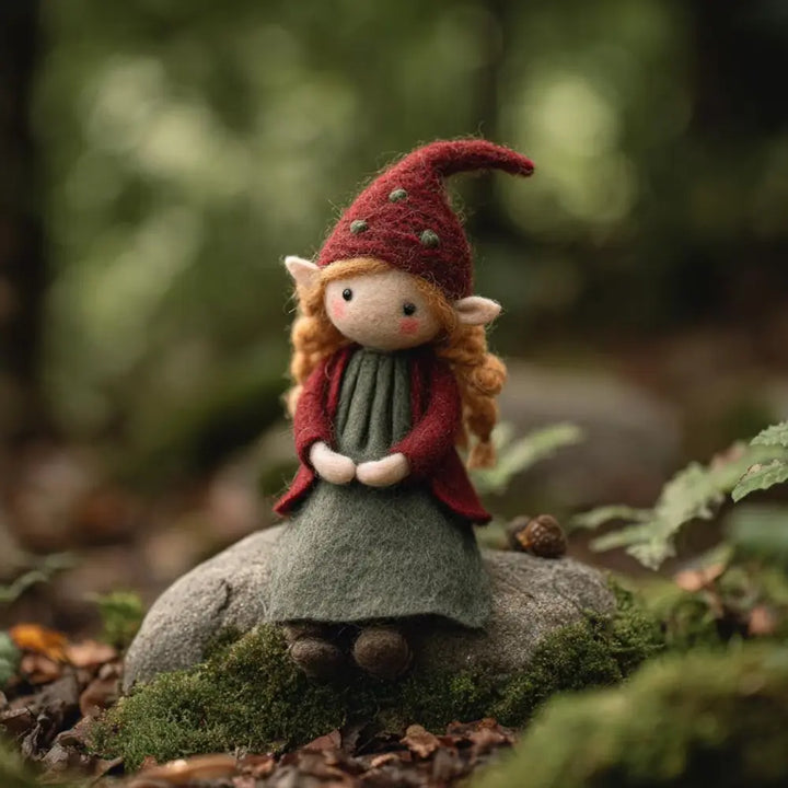 Felted gnome figurine with red hat and green dress sitting on a rock in a forest setting