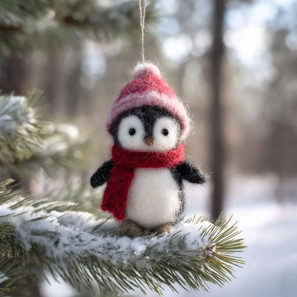 Decorative penguin ornament with a red scarf and pink hat hanging on a tree branch in a snowy forest.