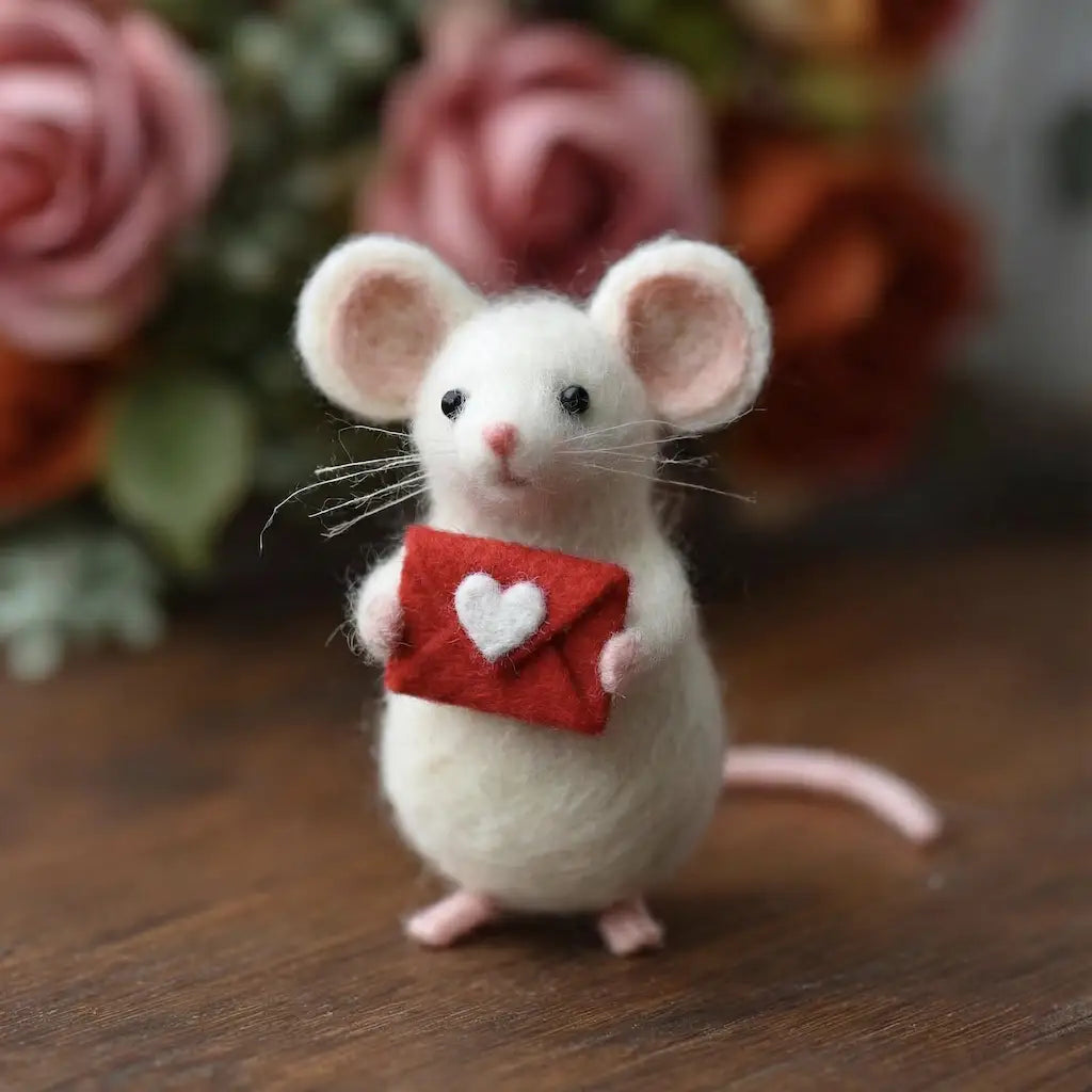 Stuffed mouse holding a red envelope with a heart on a wooden surface with flowers in the background