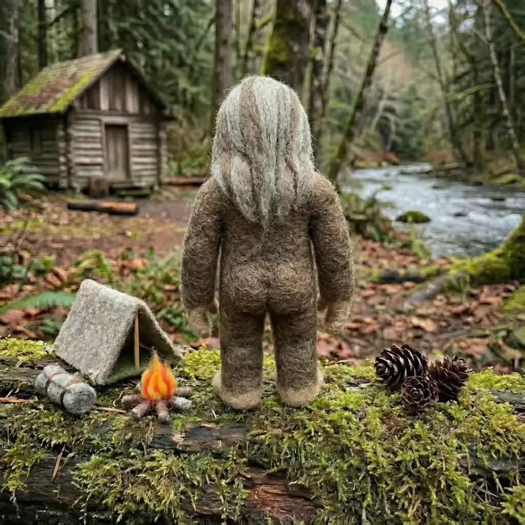 Stuffed animal resembling a bigfoot standing in a forest with a cabin and stream in the background.