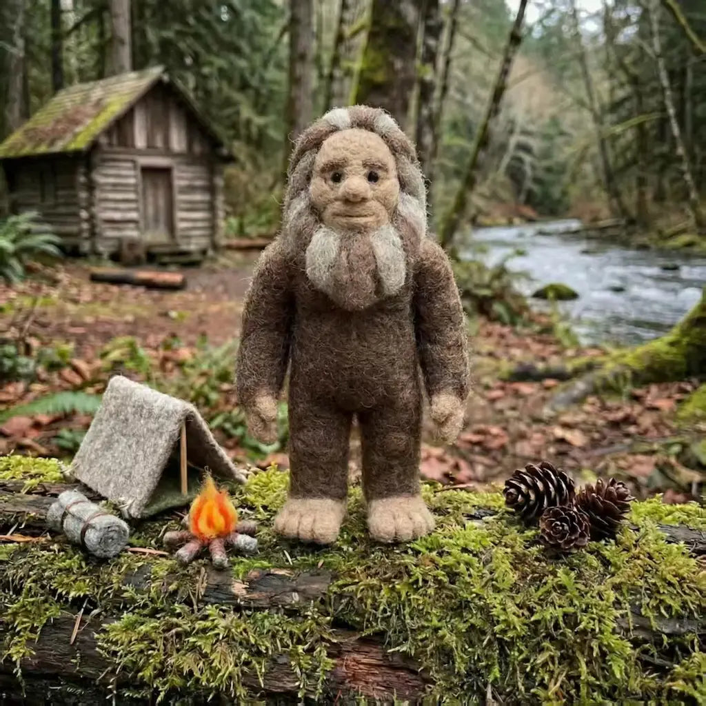 Plush toy resembling a bigfoot standing on a log with a campfire and tent in a forest setting.