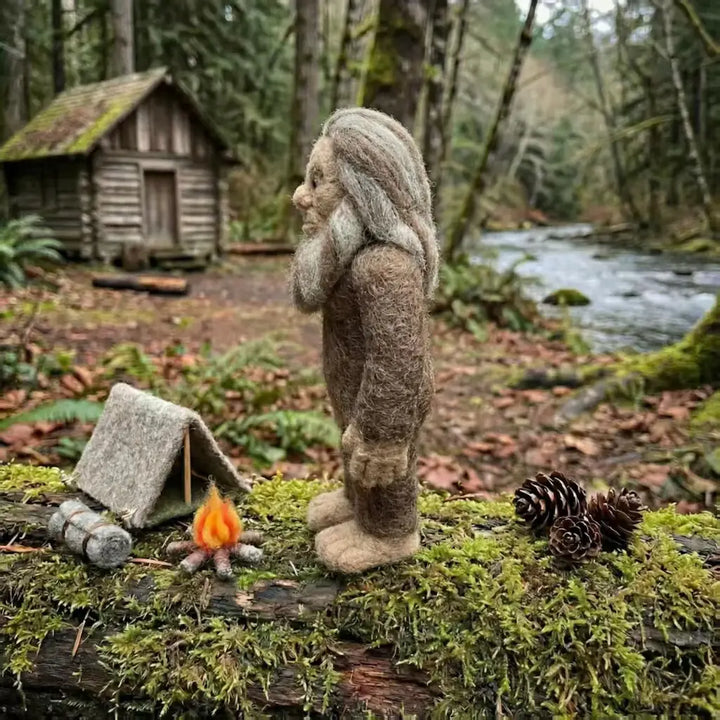 Felted gnome by a campfire and small tent in a forest setting with a cabin in the background.