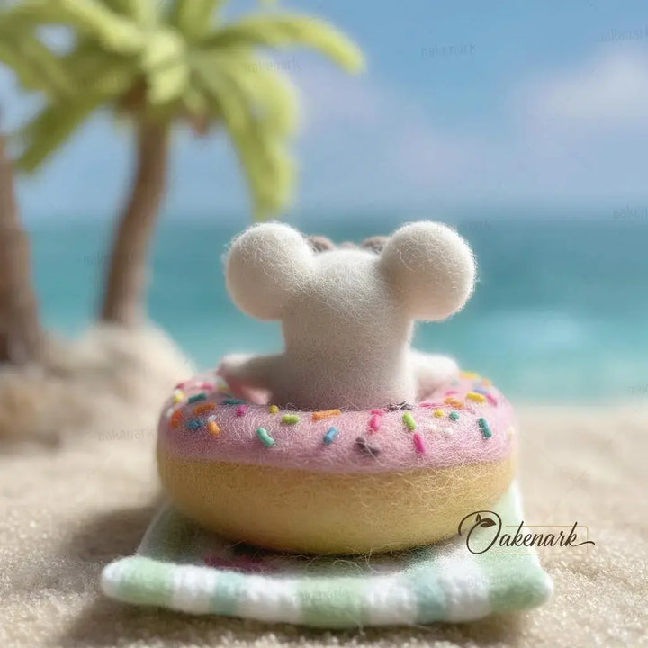 Small white bear toy on a pink donut with sprinkles on a sandy beach with palm trees and ocean in the background.