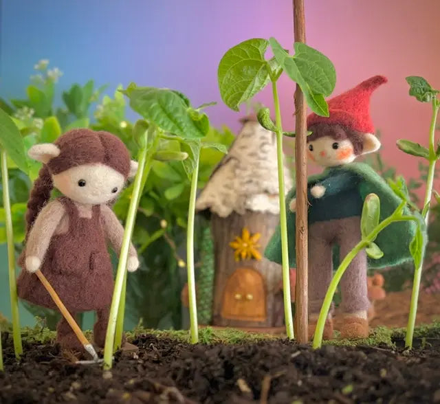 Two small figurines, one with a red hat and another with a brown outfit, standing among young plants with a miniature house in the background.