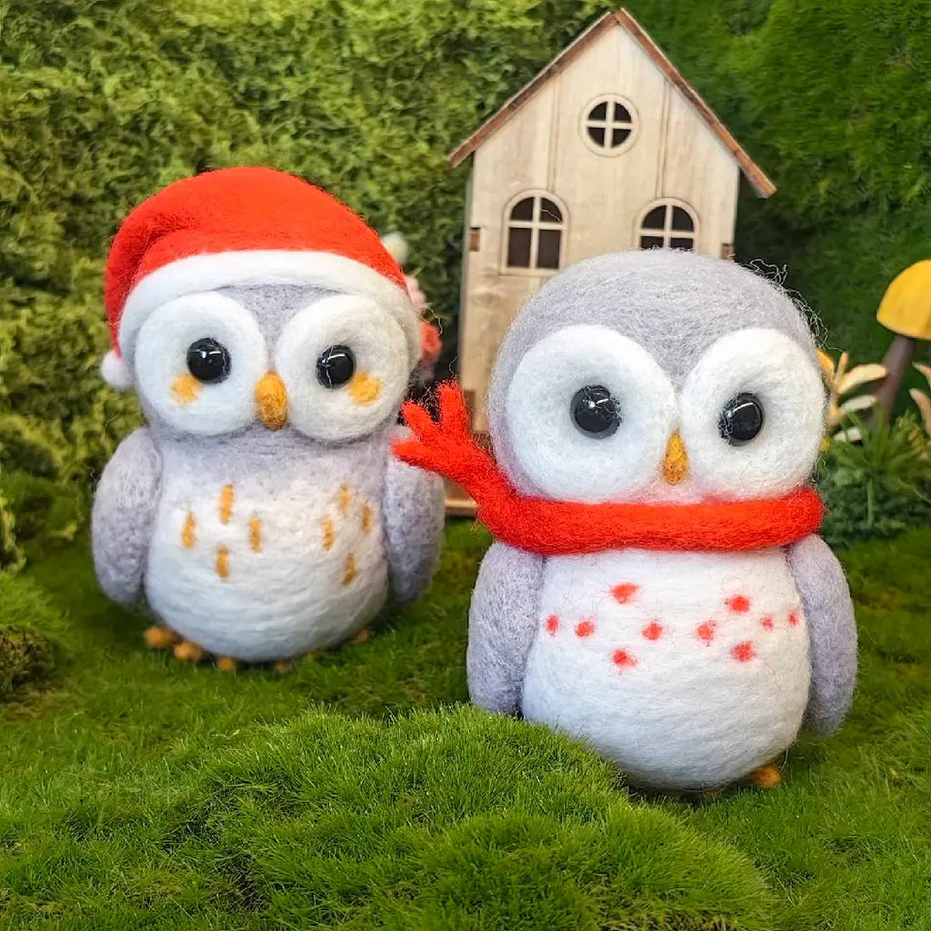 Two felt owl figurines in a mossy, forest-like setting. One owl wears a red Santa hat, and the other wears a red scarf with red spots.