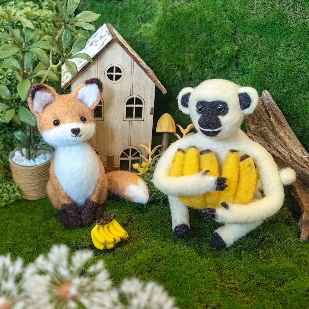 A felt wool craft display featuring a small fox and a sheep-like figure holding yellow bananas, set in a miniature garden scene with a tiny house, moss, and decorative plants.