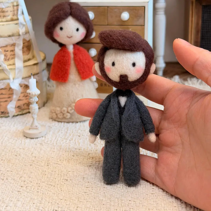 A hand holds a small felted wool doll of a man in a dark gray suit, with a second doll in a cream dress and red cardigan standing in the background on a textured beige surface.