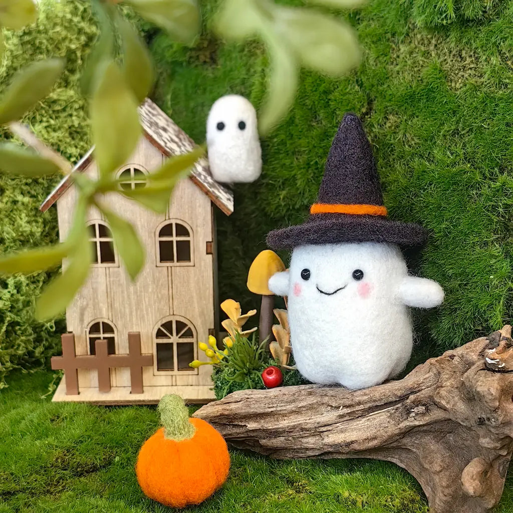 A cute white felt ghost plush toy wearing a black witch hat with an orange band, sitting next to a miniature wooden house and autumn-themed props on a mossy green surface.