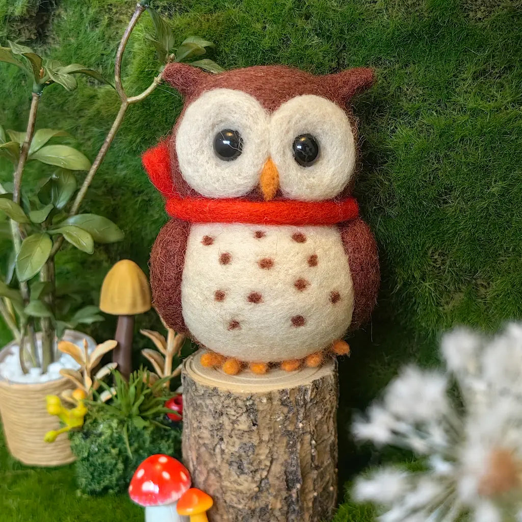 A felted wool owl decoration wearing a red scarf, perched on a small tree stump with moss, plants, and mushrooms in the background.