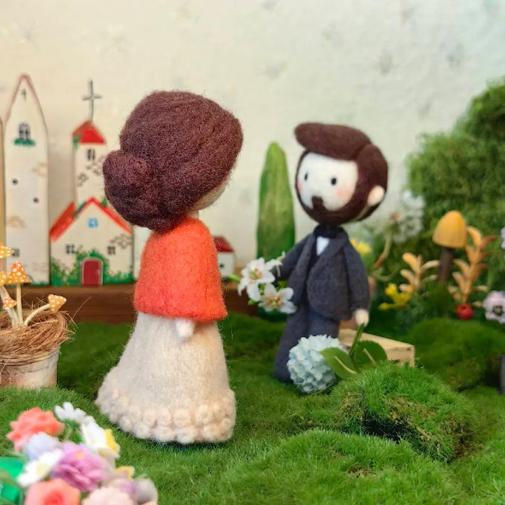 Handmade wool felt miniature figurines of a bride and groom in a tiny, decorated diorama landscape with small houses, flowers, and moss.