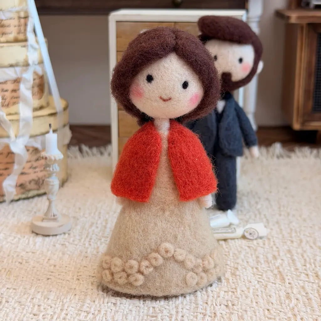 A needle-felted wedding couple figurine set on a textured cream rug. The bride figure wears a beige gown with a bright red-orange shawl, and the groom is in a dark gray suit.