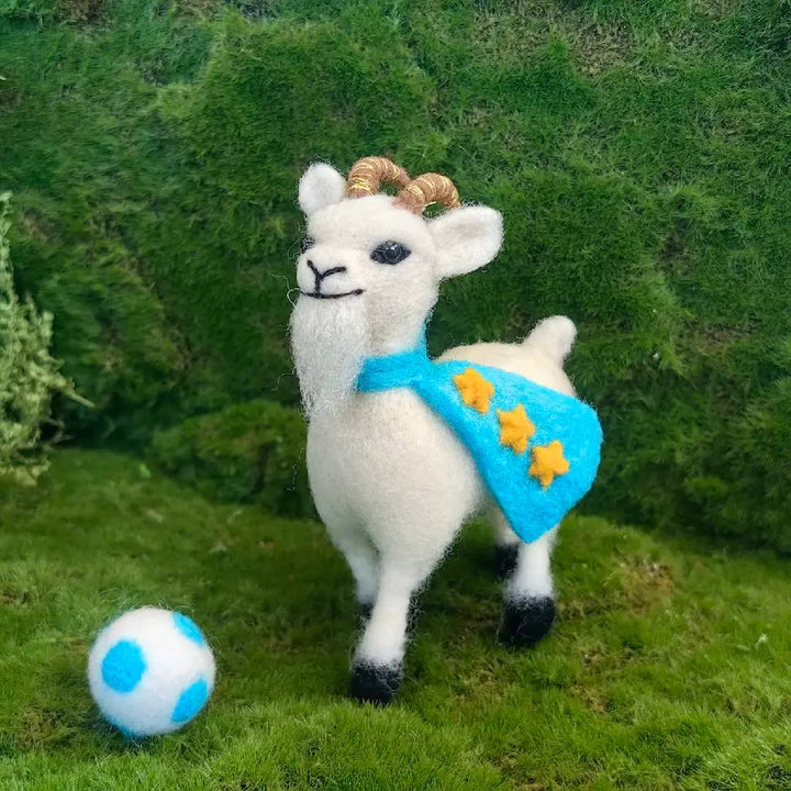 A white felted wool llama figurine with brown horns, wearing a bright blue cape with three yellow stars, standing on a green mossy surface next to a blue and white soccer ball.