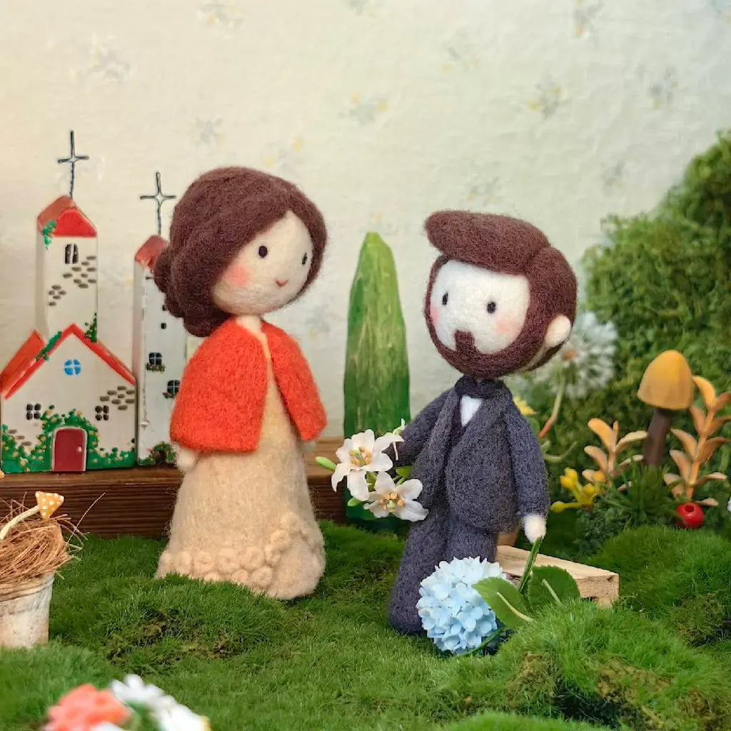 Handmade felt wool figurines of a bride and groom standing in a miniature garden diorama. The bride has brown hair, a red cape, and a cream dress, while the groom wears a dark gray suit and holds white flowers.