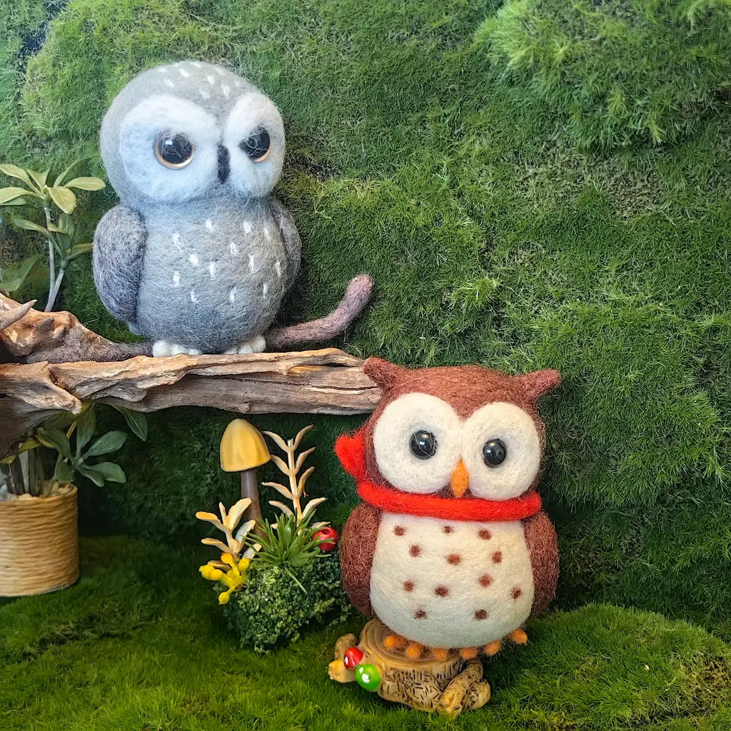 Two felt owl figurines styled in a forest scene. One gray owl with white speckles sits on a branch, and the other brown owl with a red scarf and cream spotted belly stands on a small log in front of a mossy green backdrop.