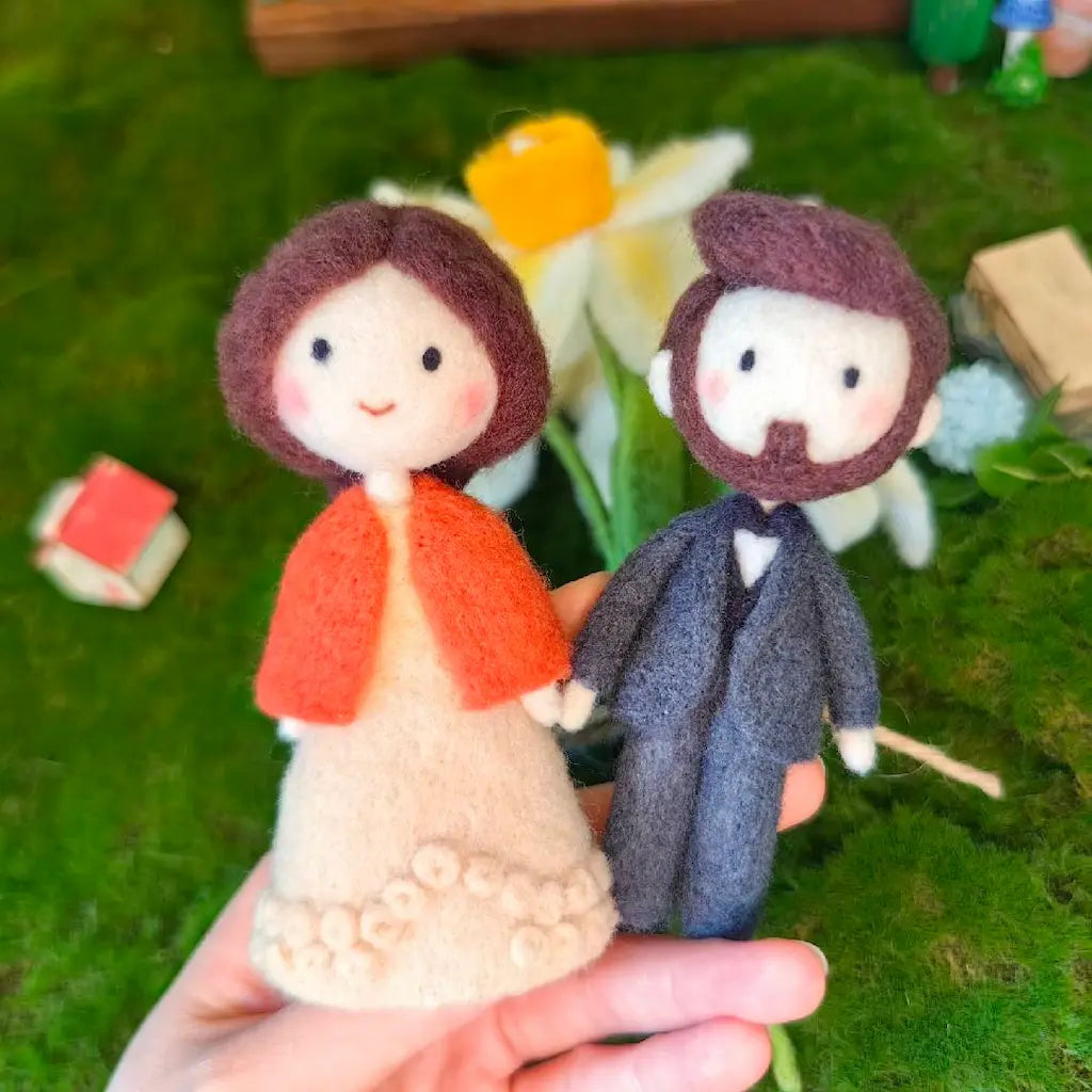 A hand holds two small felt wedding couple figurines in front of a mossy green background with blurred flowers. The bride figure wears a cream dress and orange shawl, and the groom figure wears a dark gray suit with a brown beard.