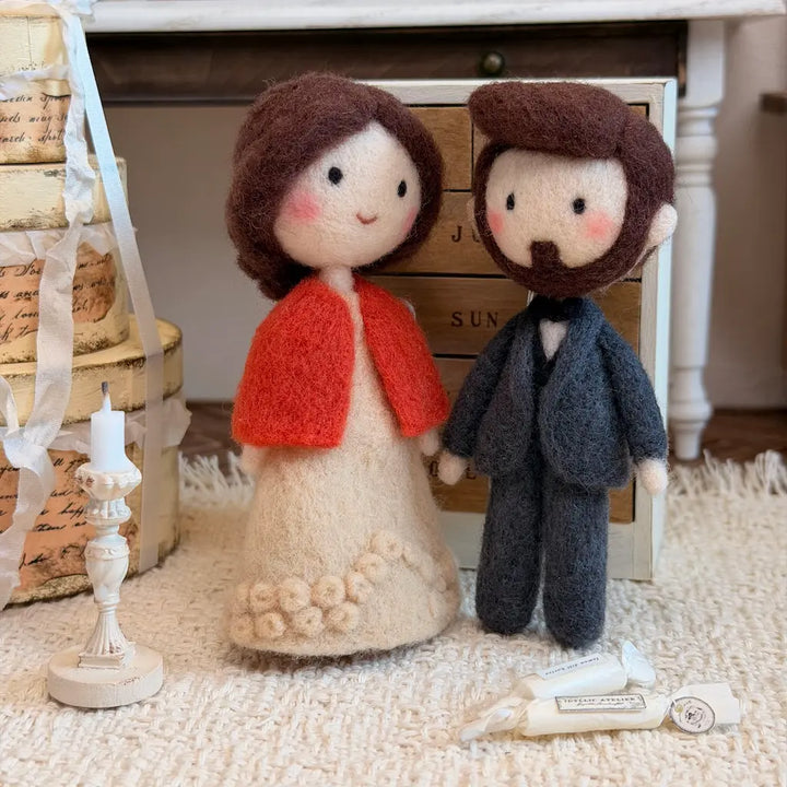 A pair of handcrafted felt wedding couple figurines, featuring a bride with brown hair and a red cape dress, and a groom with brown hair and a gray suit, posed on a white textured rug with wedding-themed props.