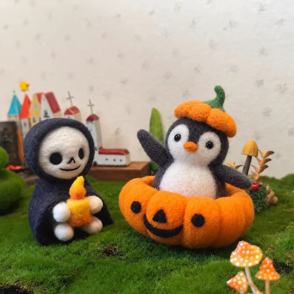 Pumpkin Parade penguin beside a hooded skull figurine holding a felt candle, arranged on moss with miniature mushrooms.