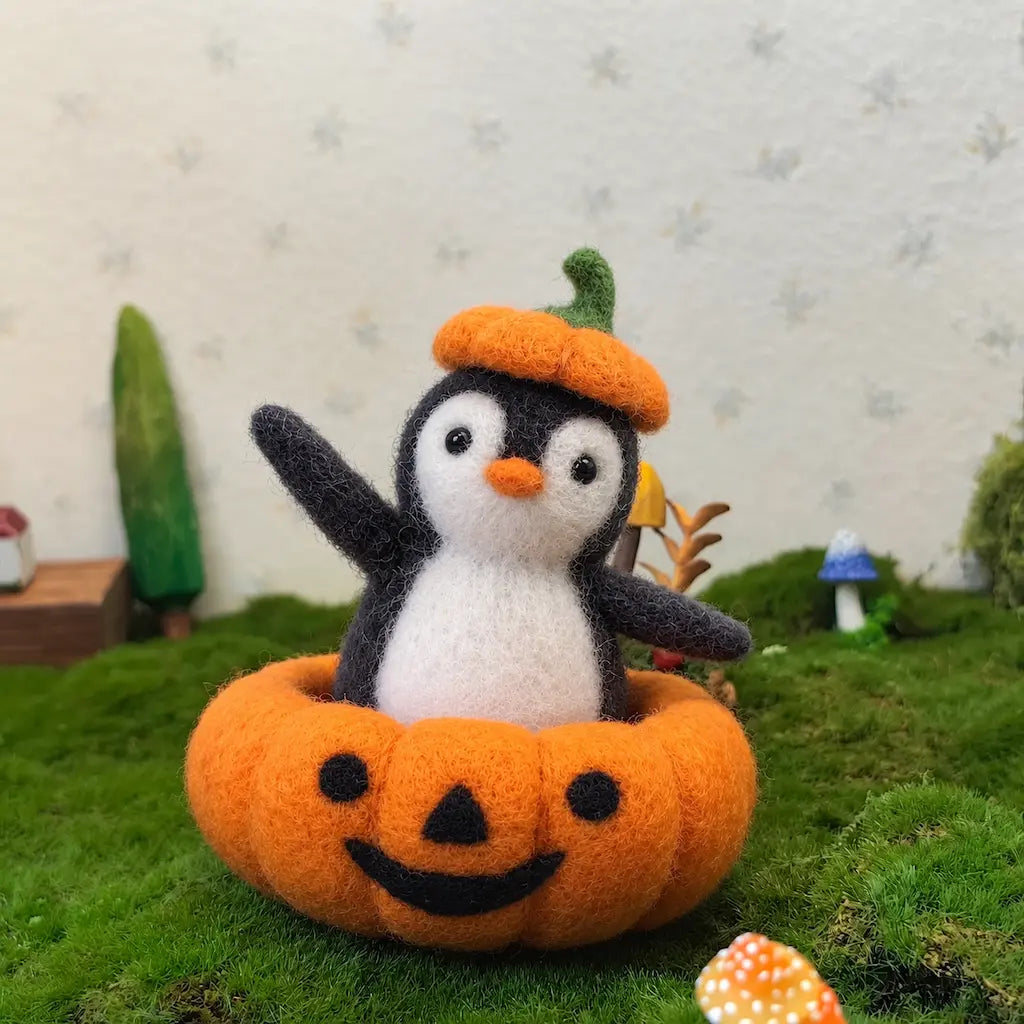 Wool felt penguin in a pumpkin costume on moss, with miniature trees and mushrooms in the background.