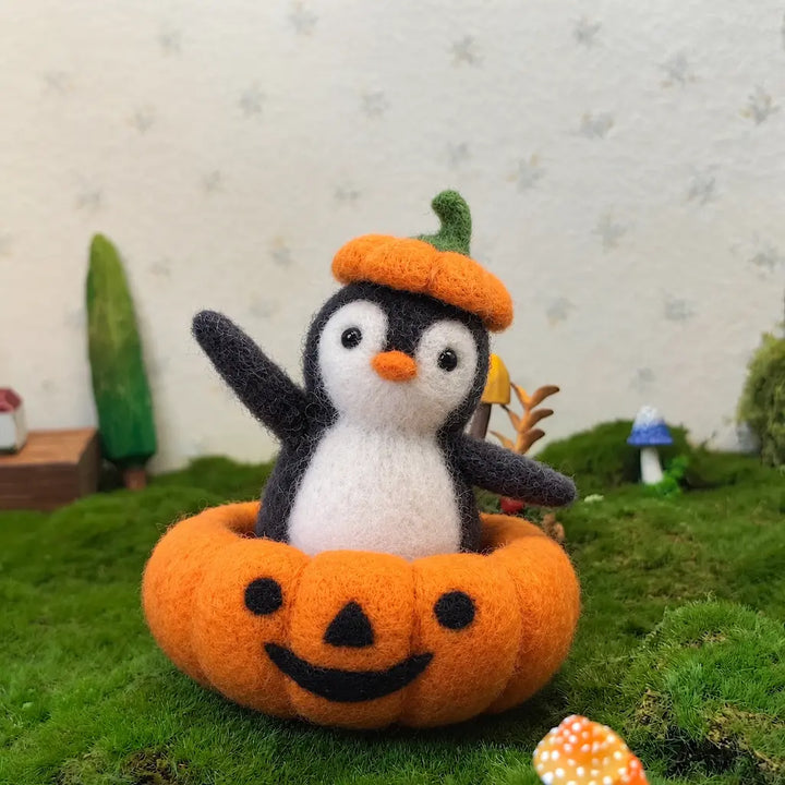 Wool felt penguin in a pumpkin costume on moss, with miniature trees and mushrooms in the background.