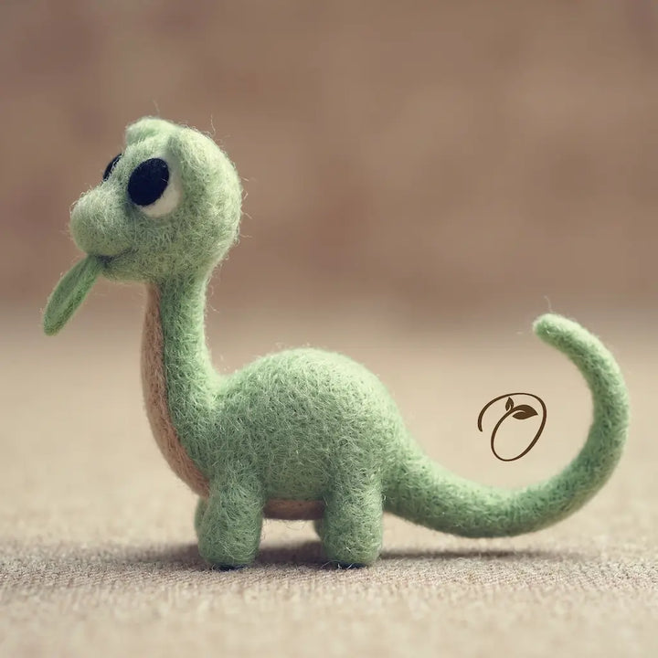 A small felt dinosaur figurine in light green with a tan underside, black eyes, and a curled tail, standing on a soft beige surface against a blurred warm brown background.