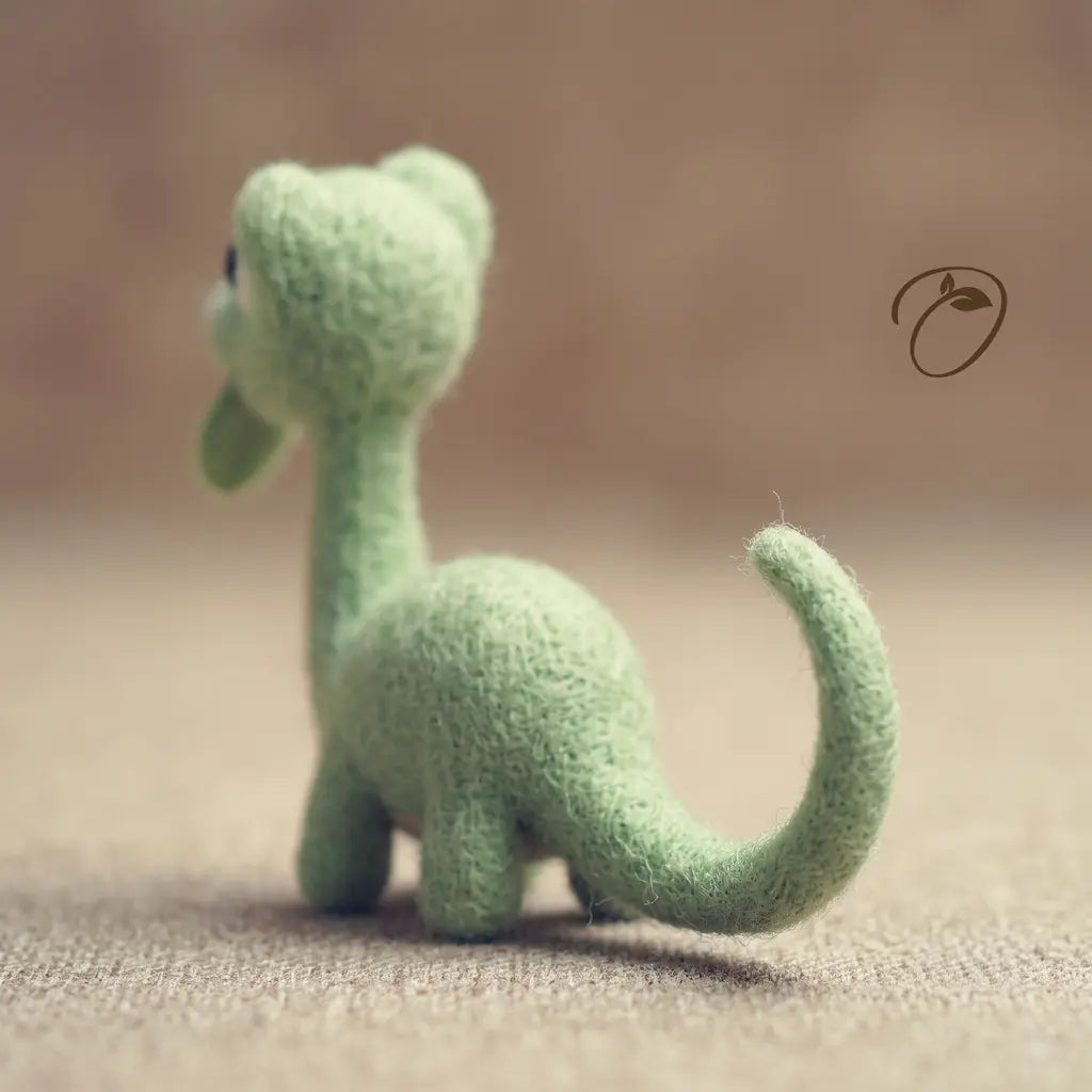 A small felt dinosaur figurine in light green, shown from the back on a soft beige textured surface with a blurred warm brown background.