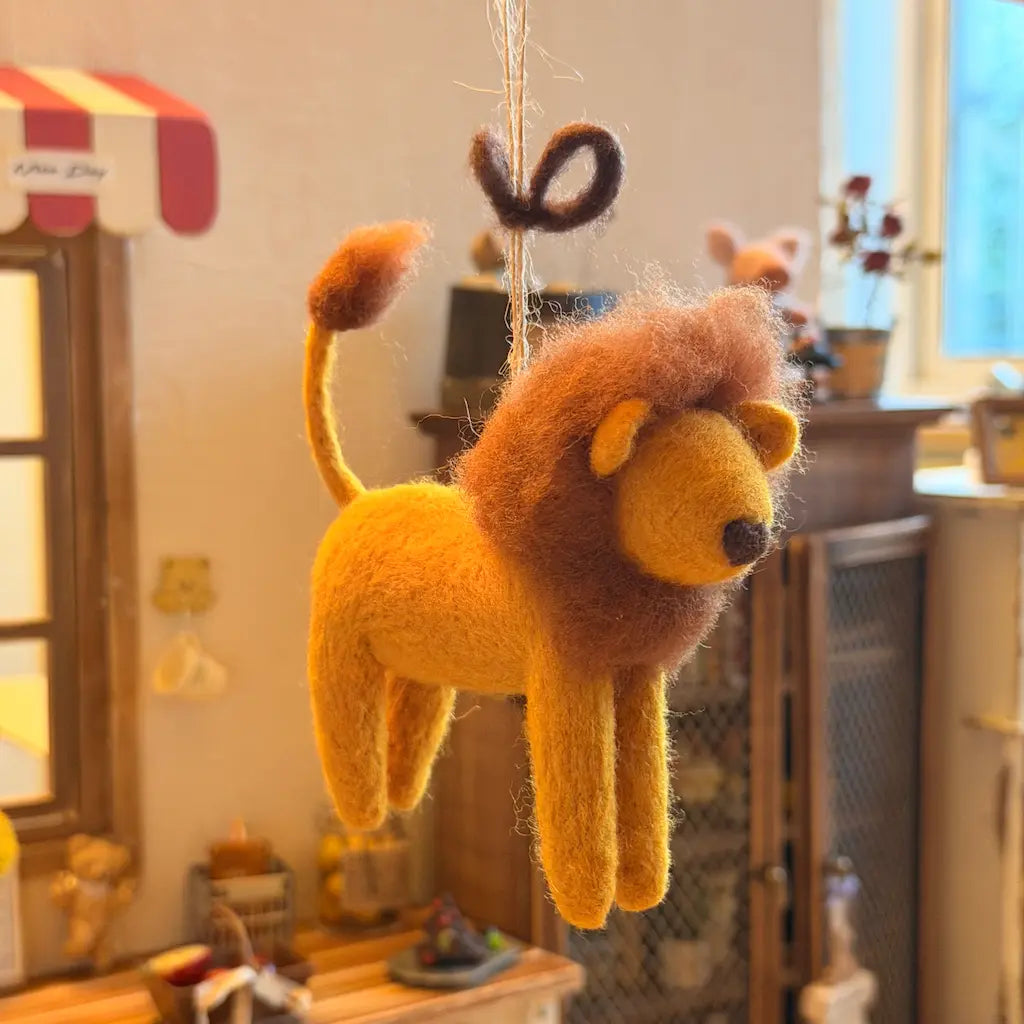 A felted wool lion hanging ornament in warm golden yellow and brown, suspended by a string in a cozy, softly blurred indoor room setting.