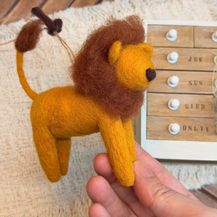 A hand holds a small felted wool lion ornament or figurine with a golden yellow body and brown mane, set against a cozy beige textured background with a wooden drawer box partially visible.
