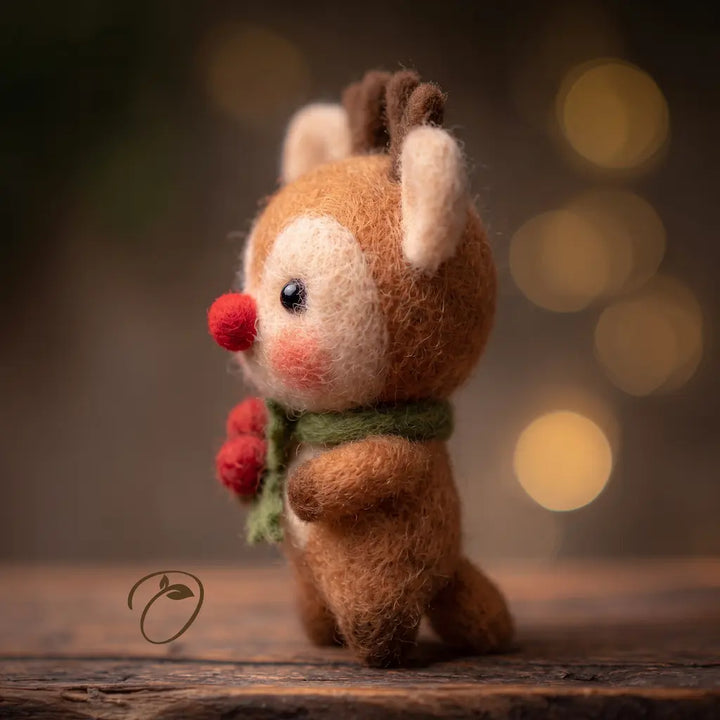 A small felted wool deer figurine with brown fur, cream face and ears, a red nose, and a green scarf with red berries, standing on a wooden surface against a warm blurred background.