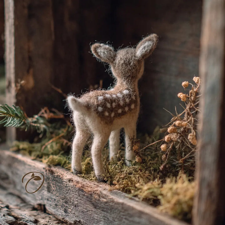A small felted wool baby deer figurine with white spots, standing on a mossy surface inside a rustic wooden display box.