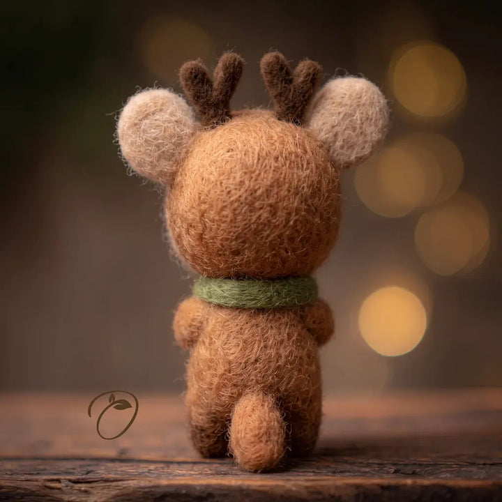 A small felted wool reindeer toy viewed from behind, sitting on a rustic wooden surface with a soft blurred warm background and bokeh lights.