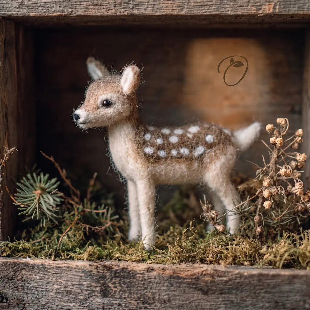 A handcrafted felted wool baby deer fawn figurine with white spots, standing on a bed of faux moss and dried botanicals inside a rustic wooden box.