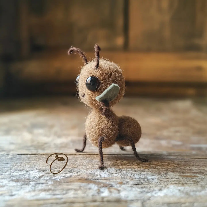 A small handmade felt ant figurine made from brown wool, featuring large black bead eyes, brown wire legs and antennae, and a light green felt wing, standing on a rustic wooden surface.