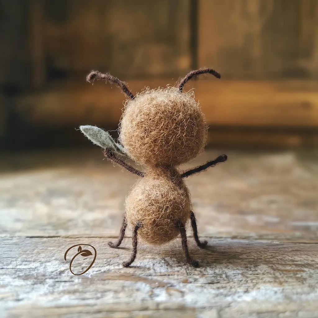 A handcrafted felt ant figurine with brown fuzzy body, dark brown antennae and legs, and a small gray-green wing, standing on a rustic wooden surface.