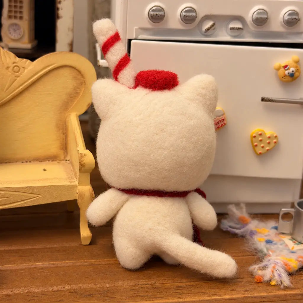Back view of the Milk Carton Cat felt figurine, showing its cream tail, red cap and striped straw beside a yellow miniature chair.