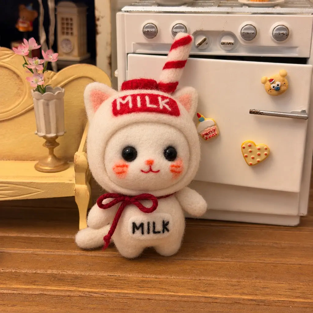 Milk Carton Cat felt figurine with a red bow and striped straw beside a yellow chair in a miniature kitchen.