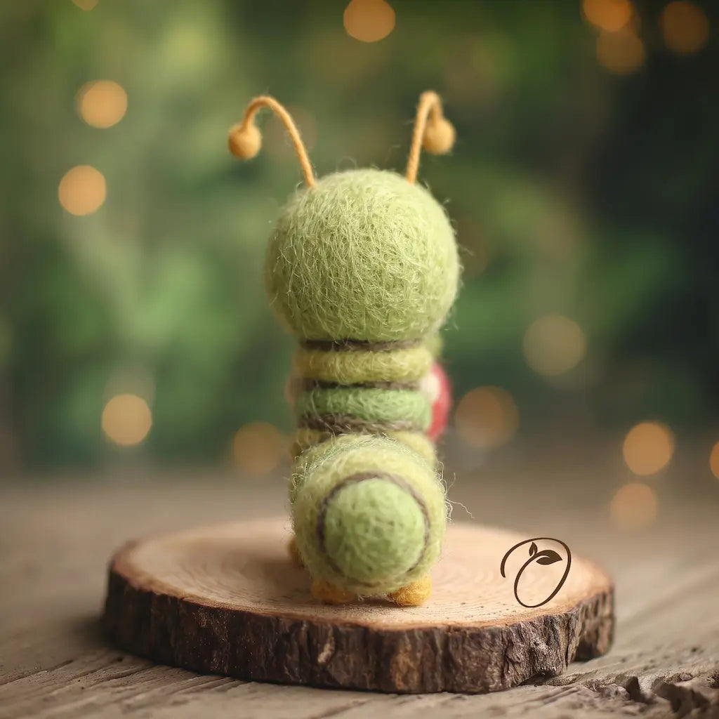 A small felted wool caterpillar figurine, shown from the back, sitting on a rustic wood slice. The caterpillar is light green with darker green stripes, yellow antennae, and a small red accent on its back.