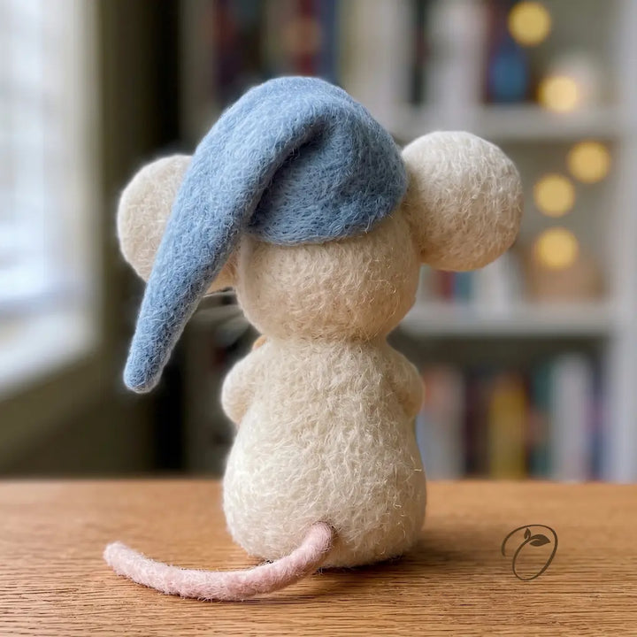 A small felted wool mouse figurine seen from the back, wearing a blue nightcap and sitting on a wooden surface with a softly blurred indoor background.