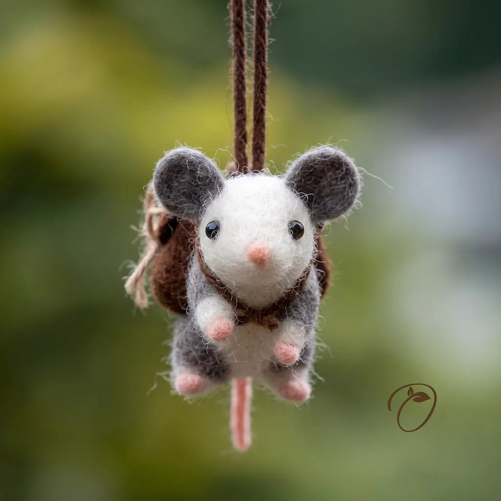 A felted wool mouse ornament wearing a small brown backpack, hanging by a brown cord against a soft blurred green outdoor background.