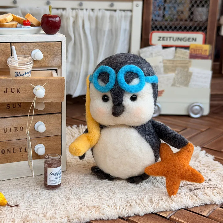 A cute felt penguin figurine wearing blue goggles and holding a yellow banana and orange starfish, sitting on a white shag rug in a miniature-style room scene.