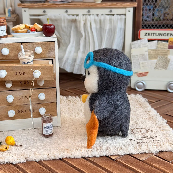 A felt penguin figurine wearing a blue aviator hat and goggles, standing on a white shag rug in a miniature dollhouse-style room with wooden floors and toy furniture.