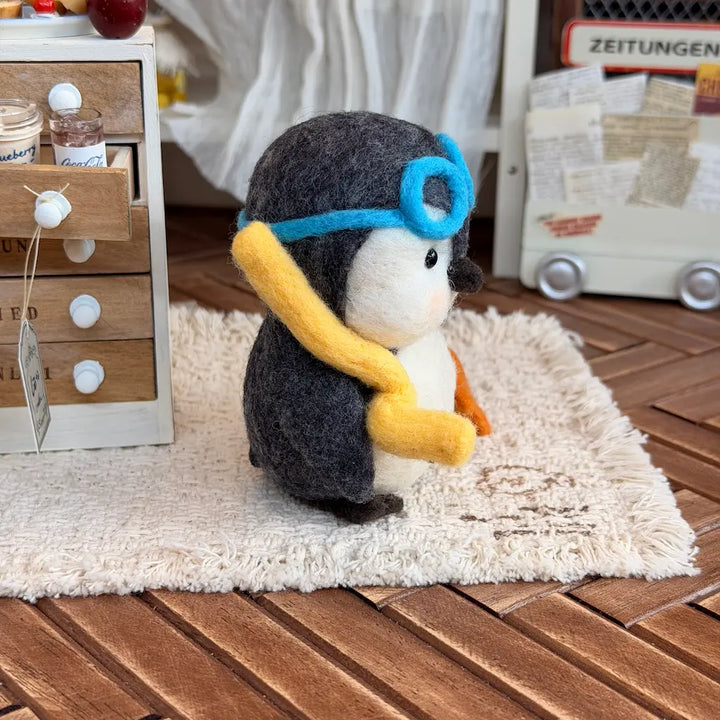 A felt penguin figurine wearing a blue goggles headband and yellow scarf, sitting on a cream shag rug over a wooden floor in a styled room setting.