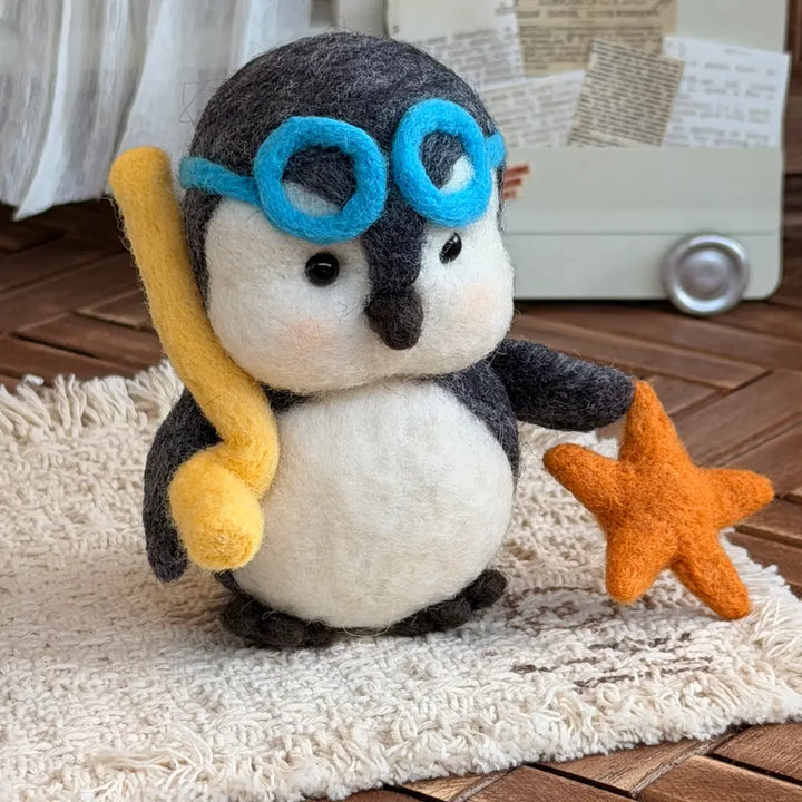 A cute needle-felted penguin figurine wearing blue goggles, holding a yellow snorkel, and standing next to an orange starfish on a cream shag rug.