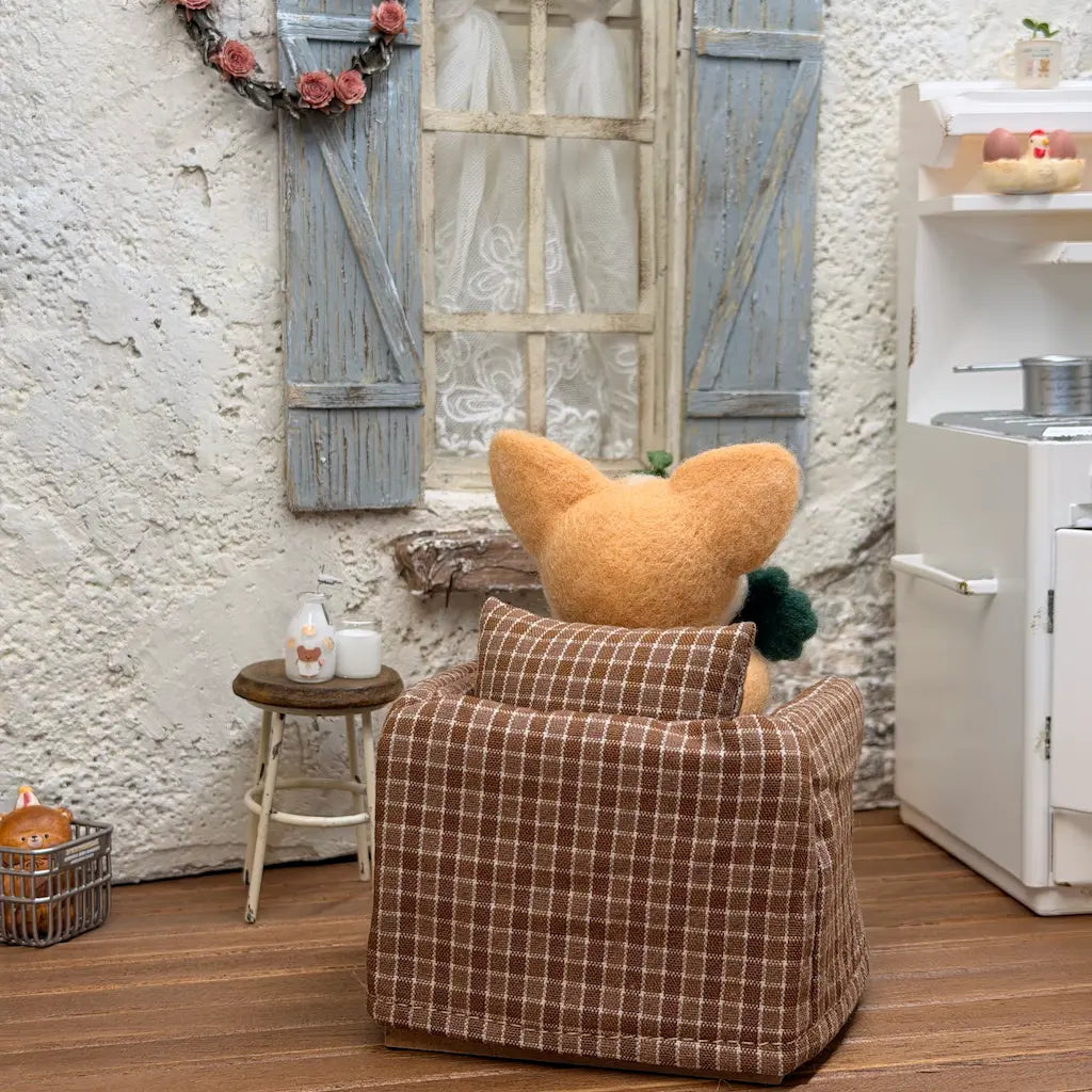 Back view of a felt corgi with a green clover in a brown checkered armchair facing a blue-shuttered window.