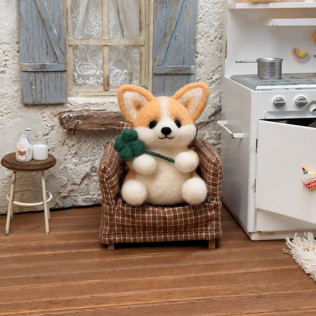 Golden tan felt corgi holding a four-leaf clover in a brown checkered armchair beside a miniature white stove.