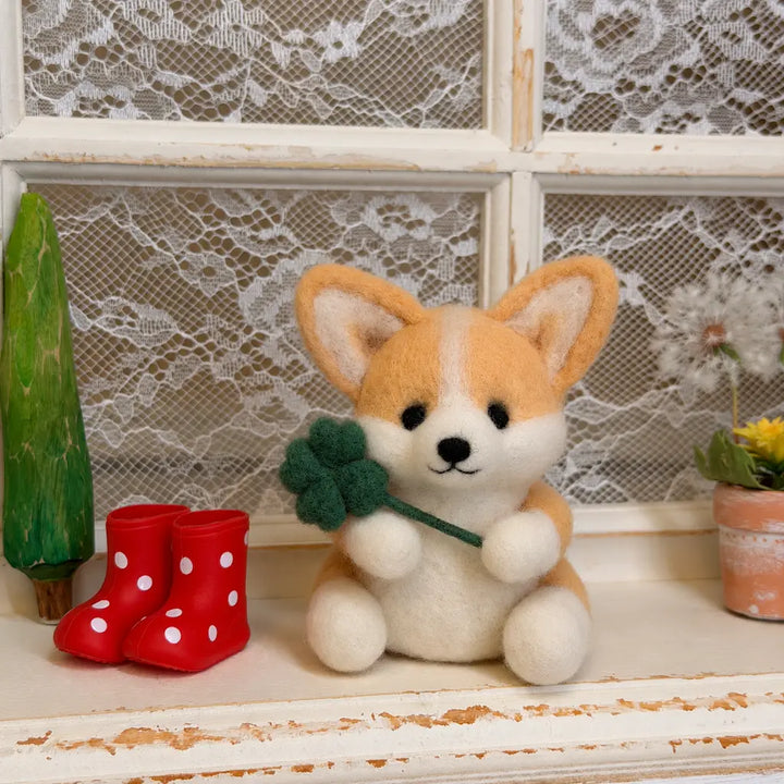 Wool felt corgi holding a four-leaf clover beside red polka-dot rain boots in front of a lace-covered window.
