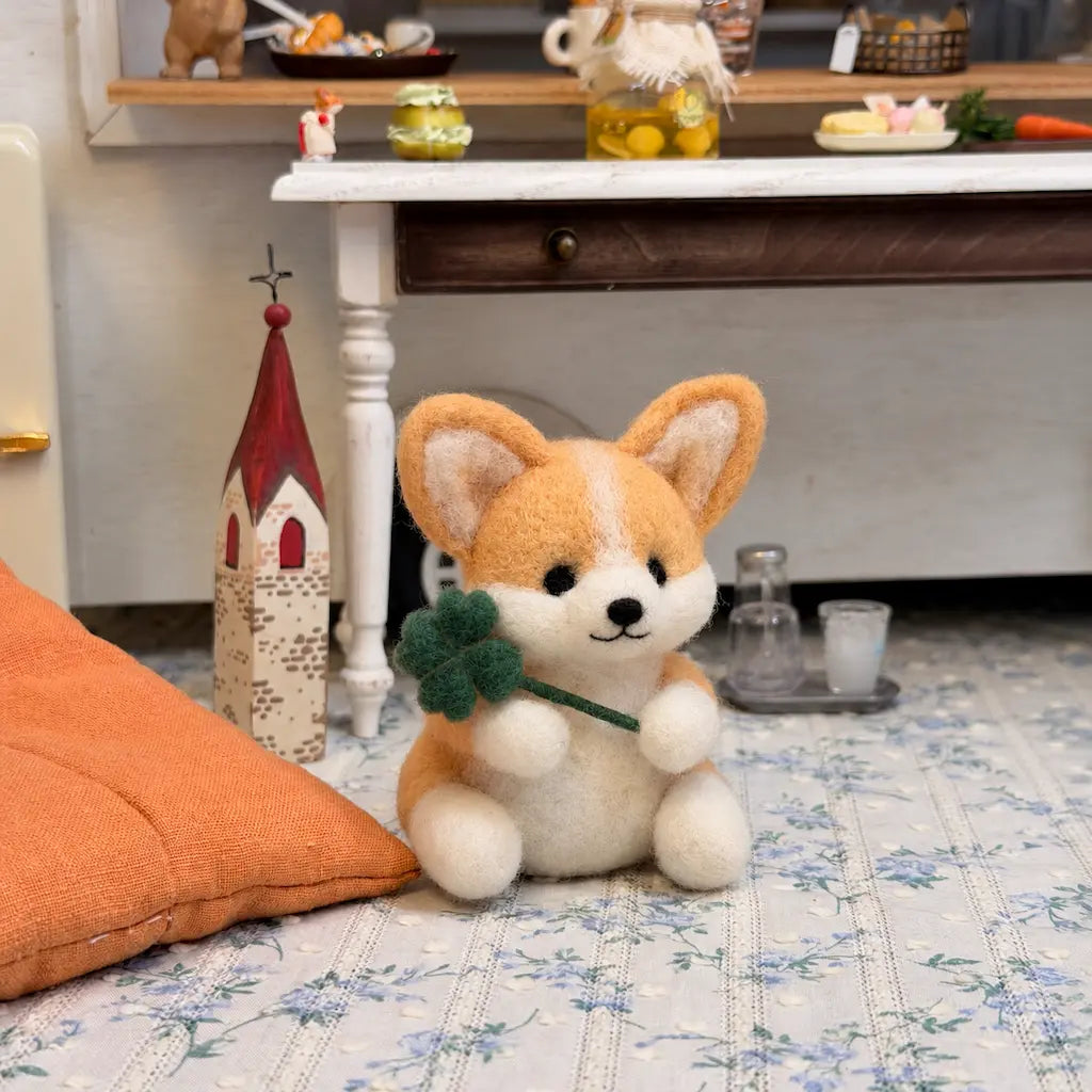 Felt corgi holding a green four-leaf clover beside an orange cushion and a miniature red-roof house.