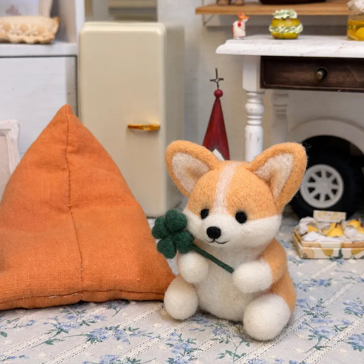 Close-up of a golden tan felt corgi holding a four-leaf clover beside a large orange cushion on floral fabric.