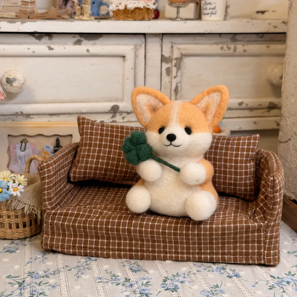 Golden tan and cream felt corgi holding a green four-leaf clover on a brown checkered miniature sofa.