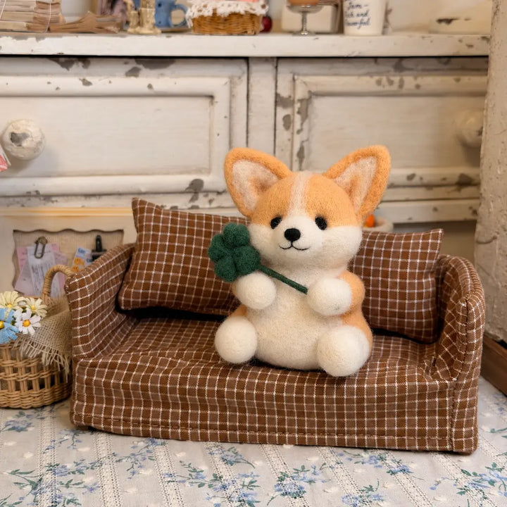 Golden tan and cream felt corgi holding a green four-leaf clover on a brown checkered miniature sofa.